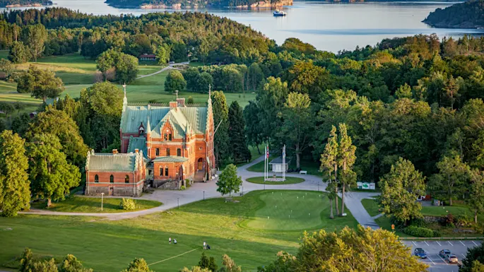 En stor, utsmykket bygning med grønt tak ligger blant frodig grønn vegetasjon, med utsikt over et rolig vann og fjerne skogkledde åser.