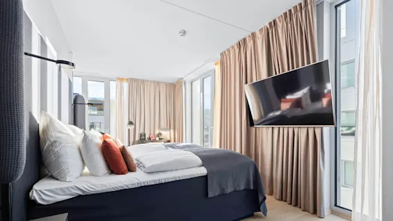A bed with multiple pillows is positioned near large windows, with beige curtains and a wall-mounted TV. Two bedside lamps are on a table, illuminating the modern bedroom.