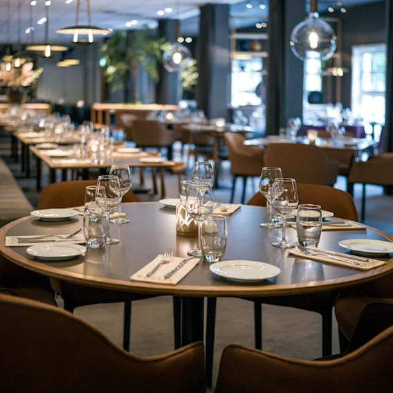 Et rundt spisebord er dekket med tallerkener, glass og servietter i en moderne restaurant, omkranset av brune stoler og opplyst av elegante pendellamper.