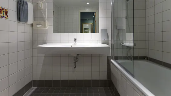 A white sink is mounted on a tiled bathroom wall, with a mirror above. A towel, hairdryer, and glass are nearby. A bathtub with a transparent screen is on the right.
