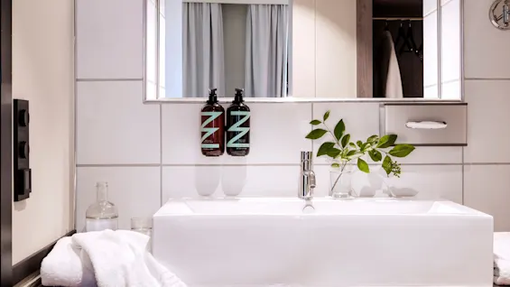 Sink with a plant-filled glass and two mounted bottles on a white tiled wall, set in a modern bathroom. A mirror reflects curtains, with towels nearby and minimalistic decor.