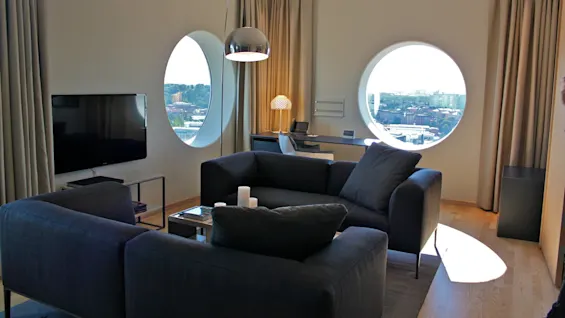 Two grey sofas face each other in a modern living room with circular windows, a wall-mounted TV, and a desk with a chair. Cityscape visible outside.