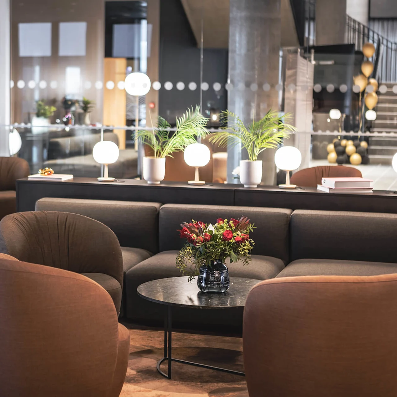 A modern lounge features comfortable brown chairs and a black sofa surrounding a round table with flowers. Soft lighting and potted plants enhance the relaxed, stylish atmosphere.