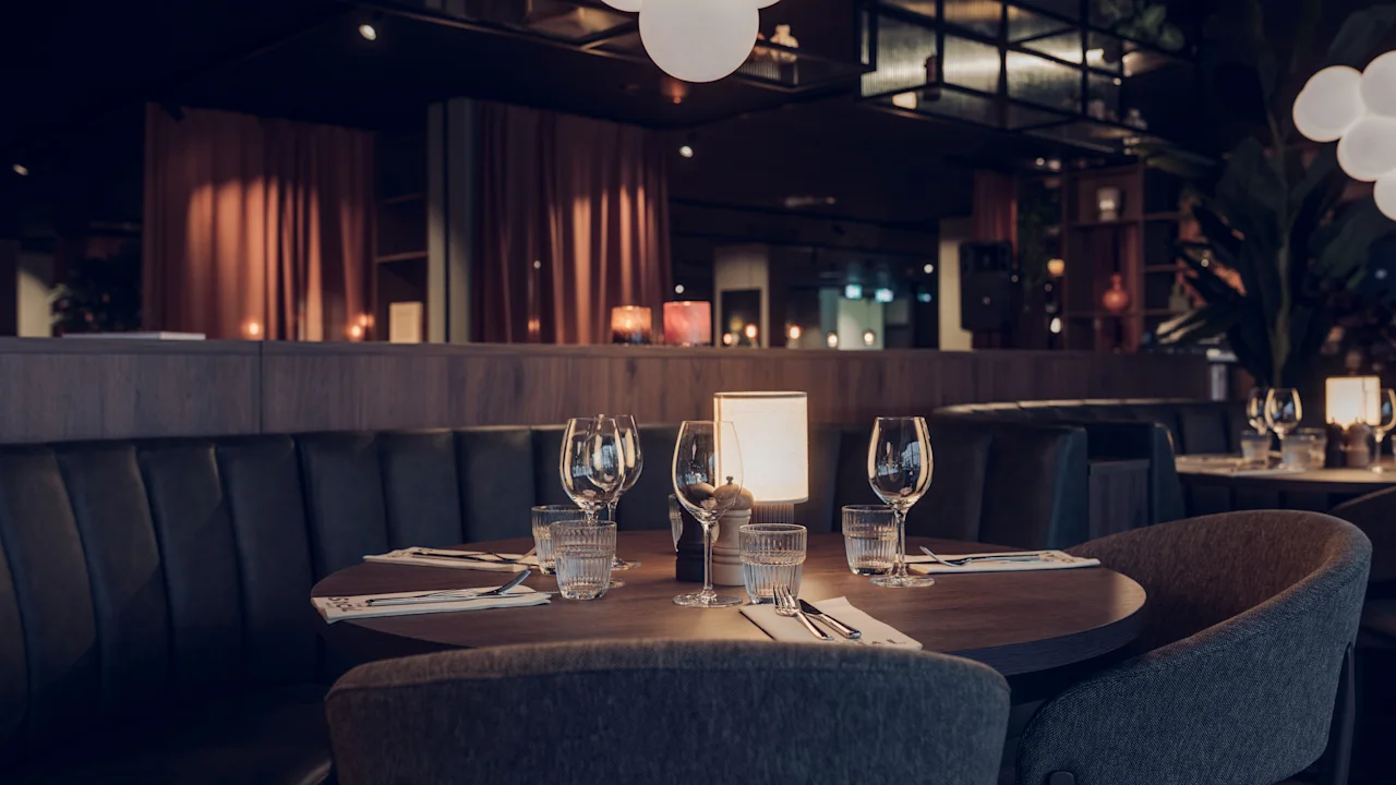 Ett runt träbord dukat med vinglas, servetter och ljus står i en svagt upplyst restaurang med mörka sittbås och mjuk belysning ovanför.