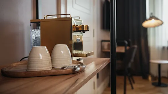 Coffee machine sits on a wooden table near two upside-down ceramic mugs on a tray; a spoon rests nearby. Soft lighting and a blurred room are in the background.