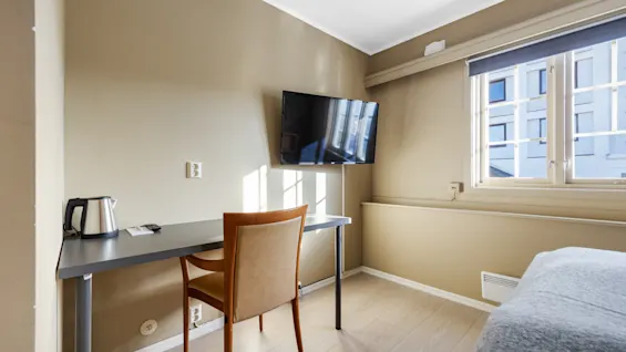 Lyst hotellrom med skrivebord, stol, veggmontert tv og et vindu som slipper inn dagslys.