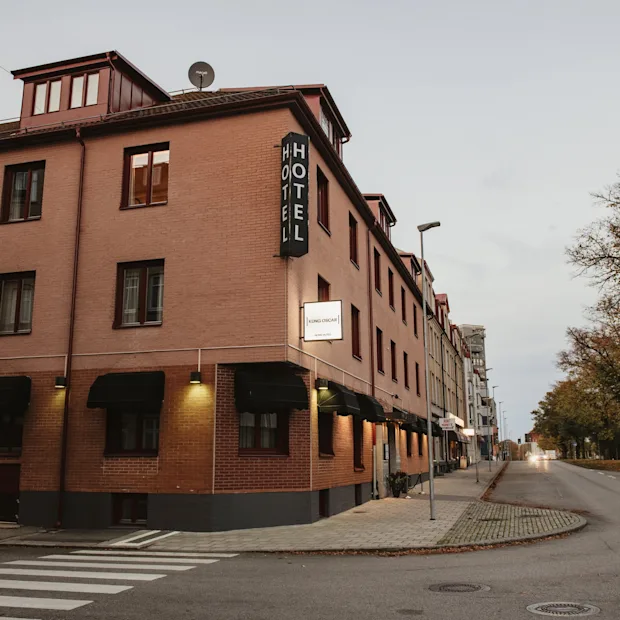A brick hotel building on a street corner with a HOTEL sign, Home Hotel Kung Oscar. Trees line the street under an overcast sky.