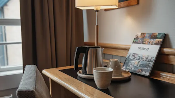 A cozy desk setup in a hotel room with a kettle, cups, and a magazine titled TROMSO, ready for a warm drink.