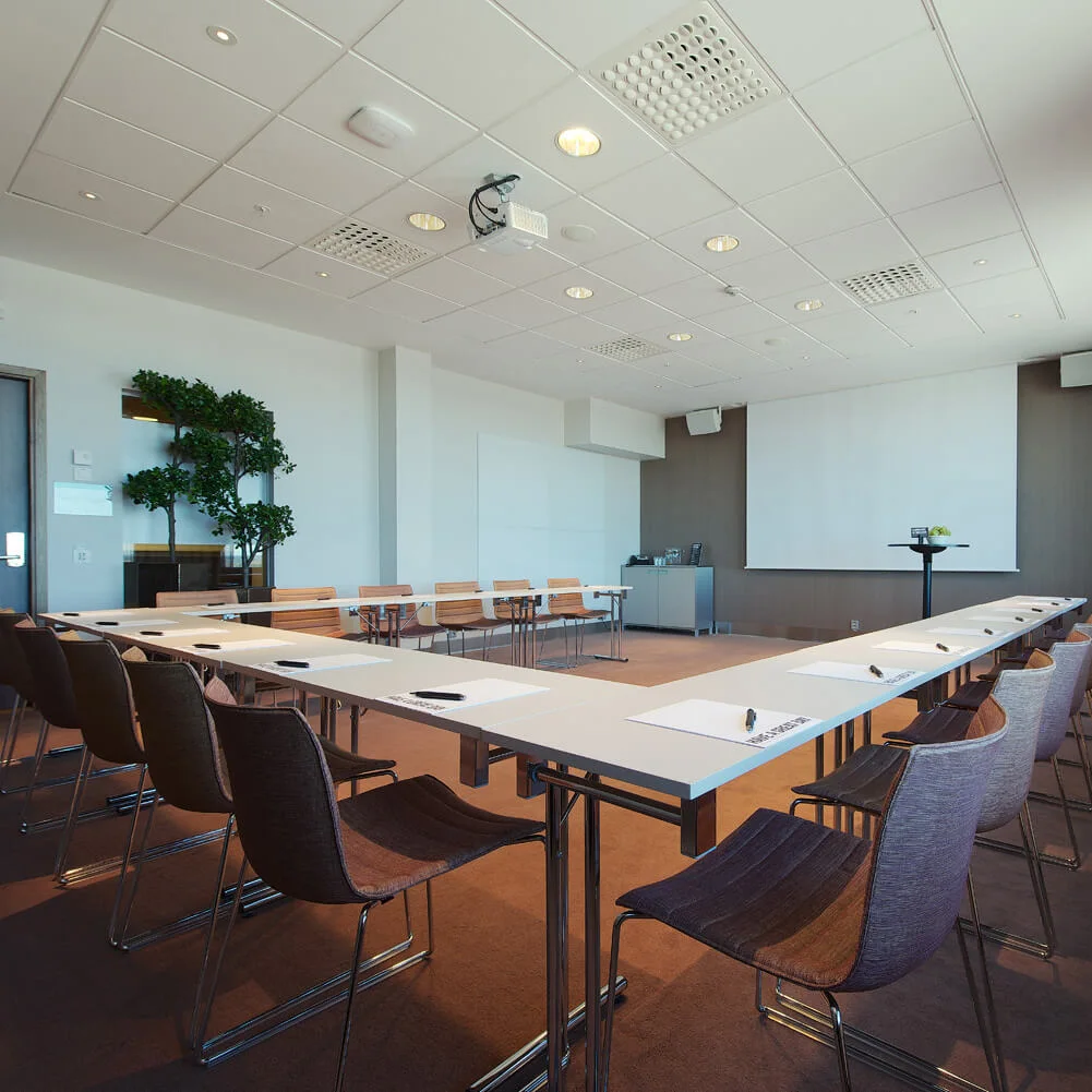 A U-shaped conference table set with notepads and pens in a bright meeting room, featuring a projector, clock, and potted plant. Large windows are covered with curtains.