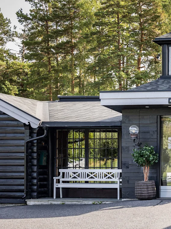 Inngang til en hotellresepsjon med glassdører, flankert av potteplanter og benker; omgitt av skogsområde. Tekst på døren: "QUALITY HOTEL ENTRY", "RESEPSJON".
