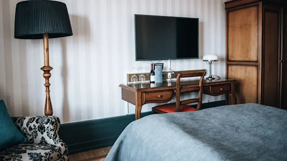 Et træbord med TV, lampe, vinflaske, glas og menu. Tæt på er der en høj lampe, blomsterstol og klædeskab, alt sammen i et smagfuldt indrettet rum.