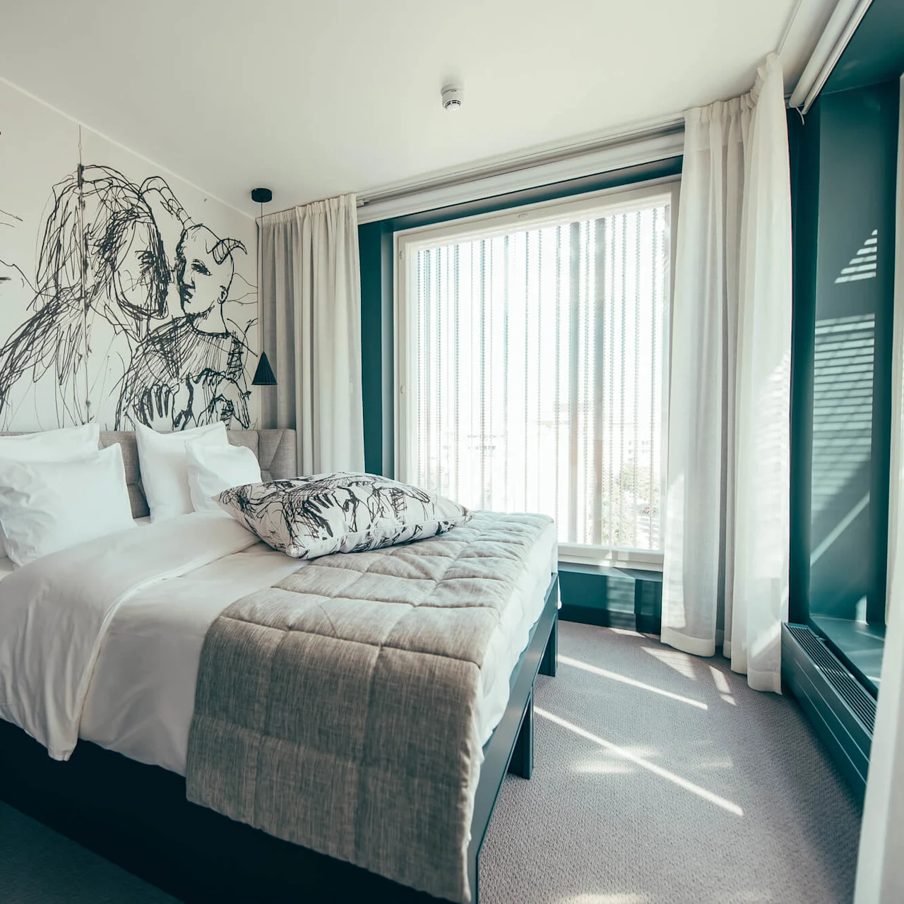 A neatly made bed with white linens and a patterned pillow rests in a room with a large mural on the wall, beside a window with sheer curtains.