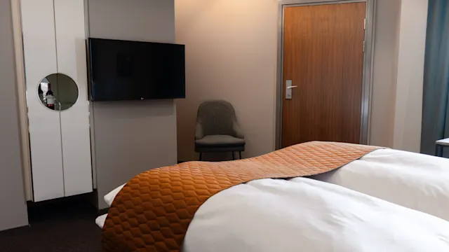 Siisti vuode, jossa on ruskea peite, hotellihuoneessa. Seinäkiinnitetty TV, suljettu puuovi ja harmaa tuoli luovat yksinkertaisen, modernin ympäristön.