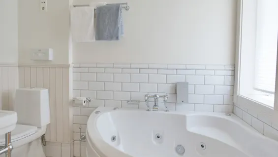 Et rent bad med et hvitt hjørneboblebad, en toalett, håndklær på en krok og t-banefliser.