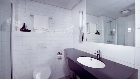 Et moderne badeværelse med hvide fliser, der har en håndvask, spejl, toilet og brusekabine.