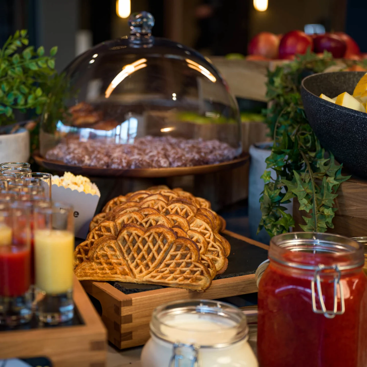 Vafler stablet pent på en treplate; omgitt av glasskrukker med sauser, en liten plante, drikkevarer, bakverk under en kuppel og frukt, alt på et buffetbord.