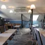 Borde med stole arrangeret i rækker, ubemandet, under skrånende loft med synlige bjælker; oplyst af hængende lamper, beliggende i et tæppebelagt konferencelokale med et sidebord tæt på et lille vindue.