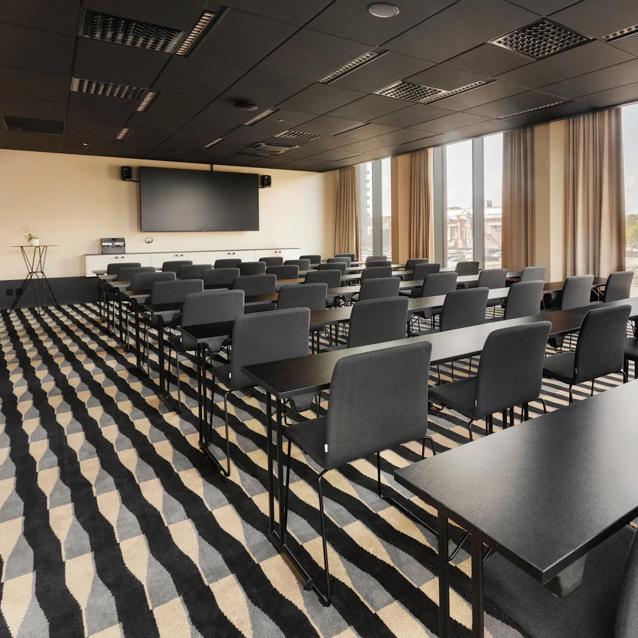 Rader av svarta stolar och bord är uppradade i ett modernt konferensrum med en stor skärm. Rummet har en mönstrad matta och stora fönster med utsikt över en stadsbild.