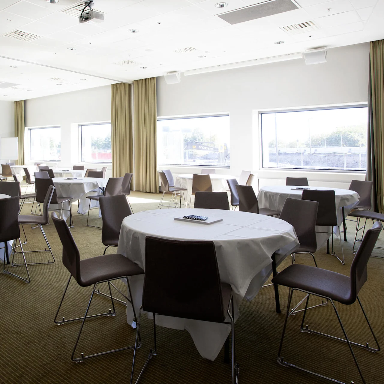 Round tables covered with white tablecloths are surrounded by brown chairs in a bright conference room. Windows allow natural light; a large screen and whiteboard are in view.