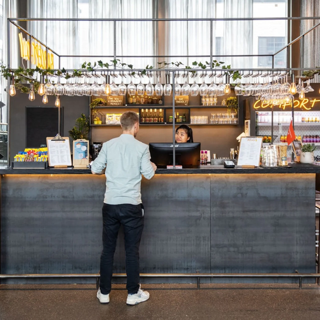 Man stands at counter in a modern café, speaking with a staff member. The counter is decorated with hanging plants and glasses; neon sign reads "COMFORT DELI."