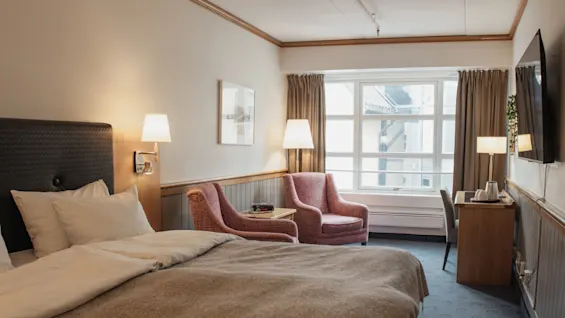 A cozy hotel room with a comfortable bed, two pink armchairs, a desk, and a large window.