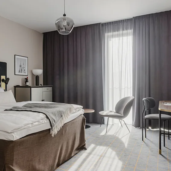 A neatly made bed with white linens is in a modern, minimalistic room with a desk, chairs, and a large window with dark curtains. A framed picture hangs above a cabinet.