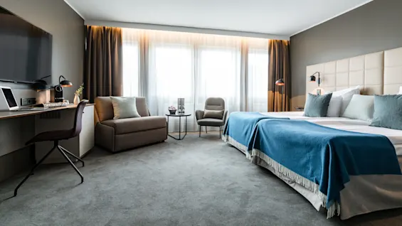 A hotel room features a neatly made bed with blue covers, a small desk with a laptop, and a cozy seating area illuminated by large windows.
