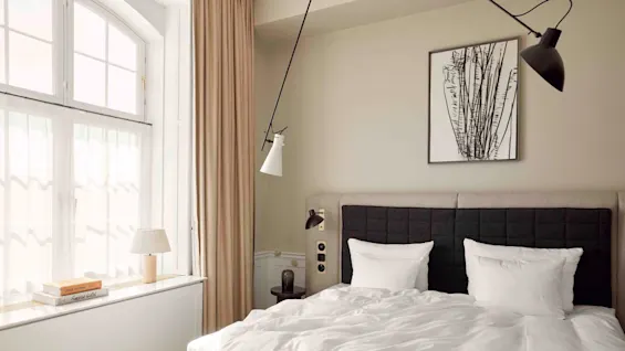A neatly made bed with white linens in a softly lit bedroom, adjacent to a window with beige curtains. A minimalist lamp and framed art decorate the space. Books titled "Louis Vuitton" and "Fangtastic Girls" are on the windowsill.
