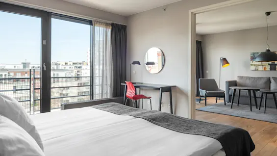 A neatly made bed with white sheets and a gray blanket faces a sliding glass door to a balcony. A red chair and desk sit beneath a round mirror, next to an adjoining living area with gray furniture.