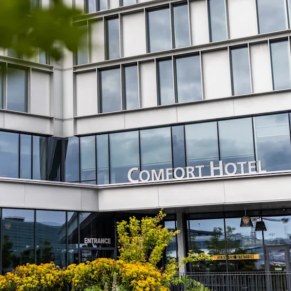 Nærbillede af et moderne hotel med store glasfacader, omgivet af grønne buske og gule blomster i forgrunden. Indgangen og hotellets skilt er synlige.
