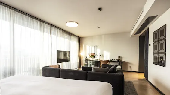 Et romslig, moderne hotellrom med en stor seng i forgrunnen, vendt mot en flatskjerm-TV. En grå sofa står i nærheten. Myk belysning kommer fra en lampe og et vindu.