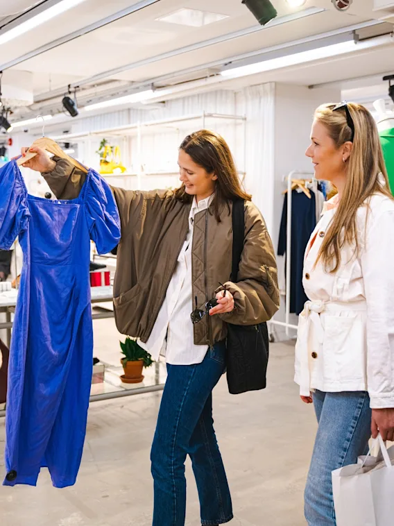 Tre kvinnor shoppar i en klädbutik, en håller upp en blå klänning för sina vänner att se.