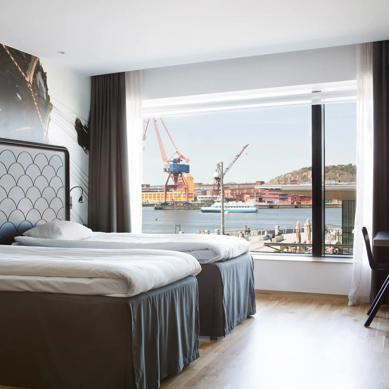 Två prydligt bäddade enkelsängar bredvid ett stort fönster med utsikt över en hamn, där kranar och ett skepp syns. Rummet har modern inredning med väggkonst.