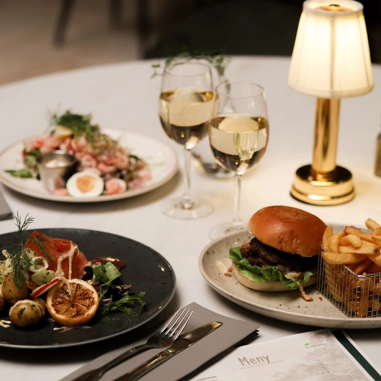 Burger with fries sits on a table next to a salad, two glasses of white wine, a plate with smoked salmon and potatoes, a menu, and a small lamp. 
