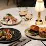 Burger with fries sits on a table next to a salad, two glasses of white wine, a plate with smoked salmon and potatoes, a menu, and a small lamp. 