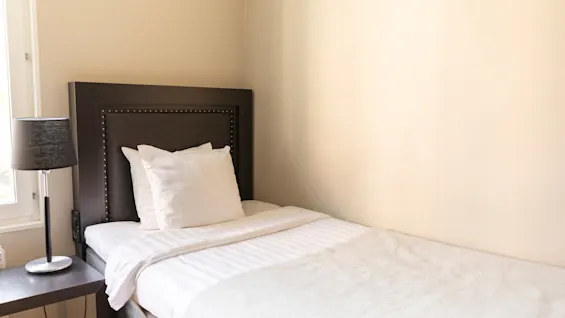 En mysig enkelsäng med vita sängkläder och en mörk sänggavel, bredvid ett sängbord med en lampa, på Home Hotel Tapto i Stockholm.