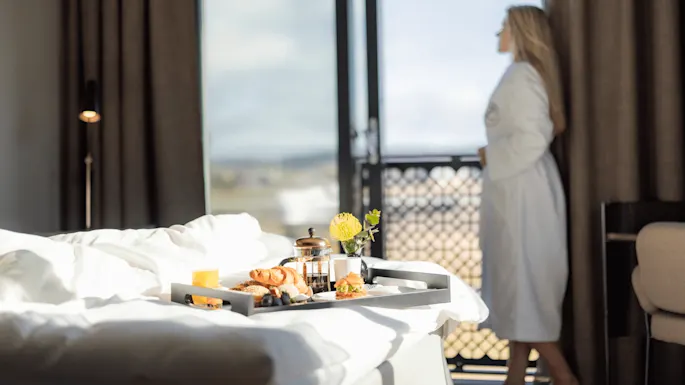 A breakfast tray on a bed with a blurred person in a bathrobe looking out of a sunny hotel room window.