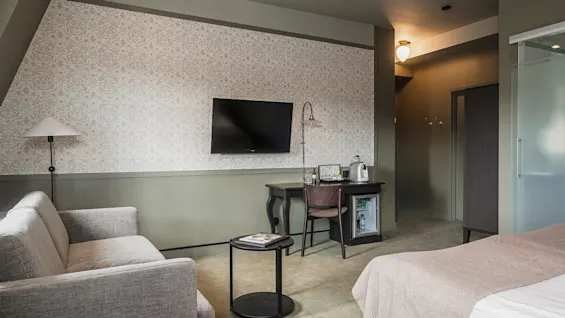 Ett hotellrum med säng, soffa, skrivbord med minibar, hänglampa, väggmonterad TV och mönstrad tapet i en mysig miljö.
