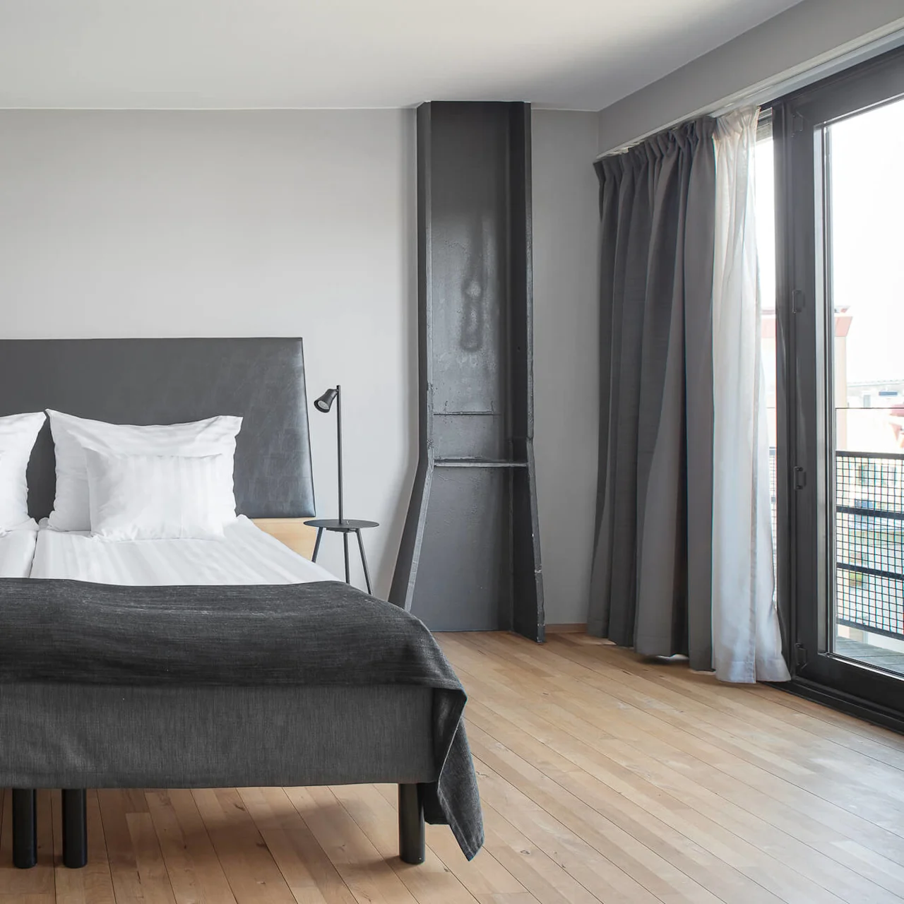 A neatly made double bed with white pillows and dark grey bedding sits in a modern bedroom with wooden floors and minimalist decor, adjacent to a window with grey curtains.