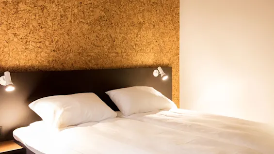 A neatly made double bed under two wall-mounted lights, situated against a textured cork wall, creates a warm and cozy atmosphere in the softly lit room.