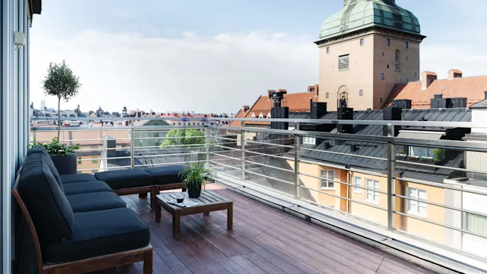 A balcony with comfy seating overlooks a cityscape with historic and modern buildings under a clear sky.
