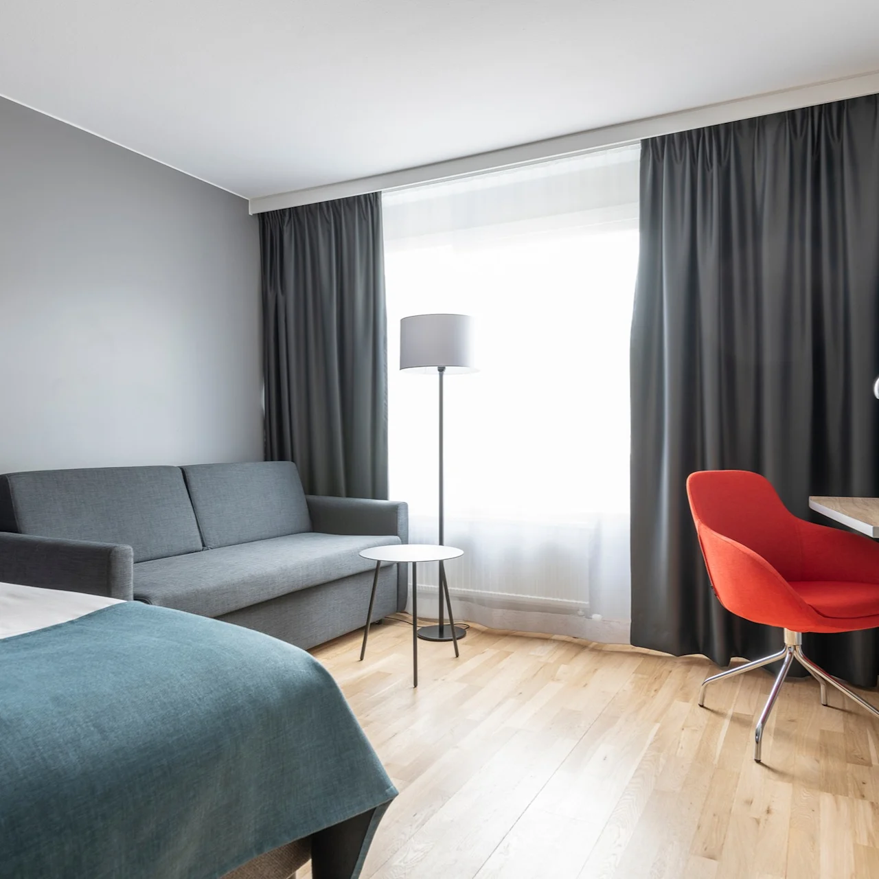 En grå soffa står i ett modernt rum med en golvlampa och litet bord vid fönstret. En orange stol är placerad vid ett skrivbord.