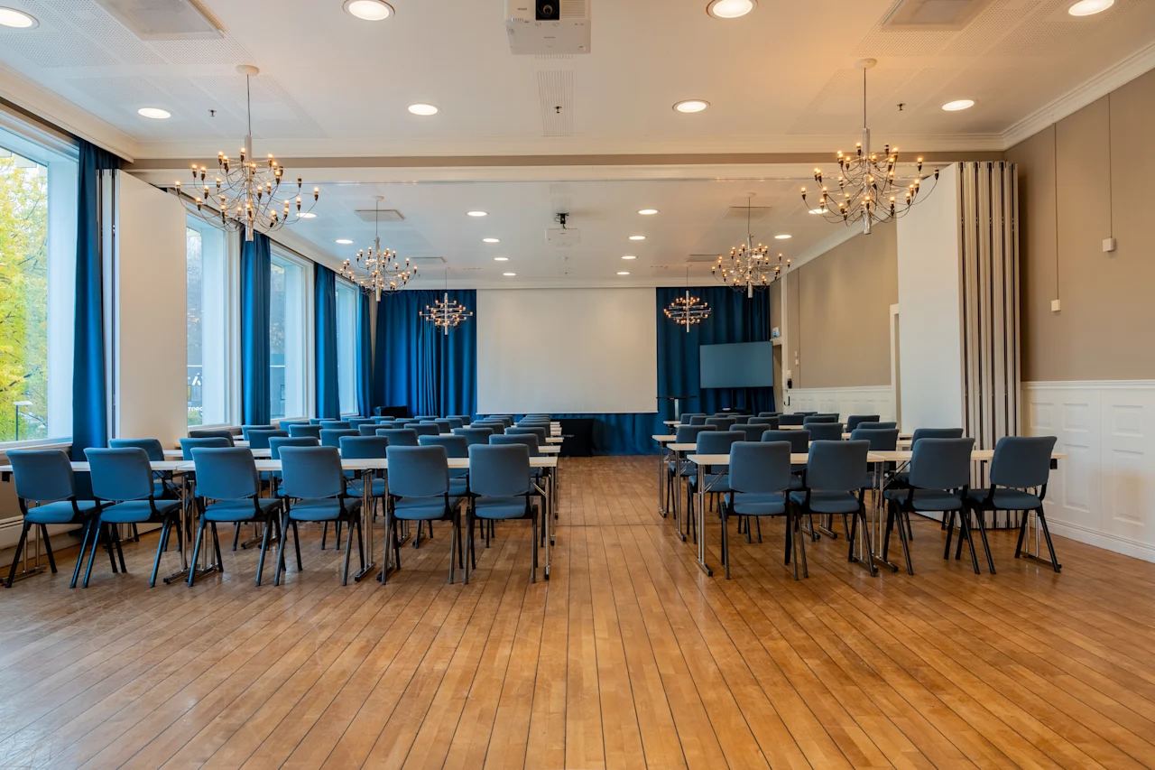 Tilava kokoushuone, jossa on riveittäin pöytiä ja tuoleja, suuri valkokangas ja ikkunat sinisillä verhoilla.
