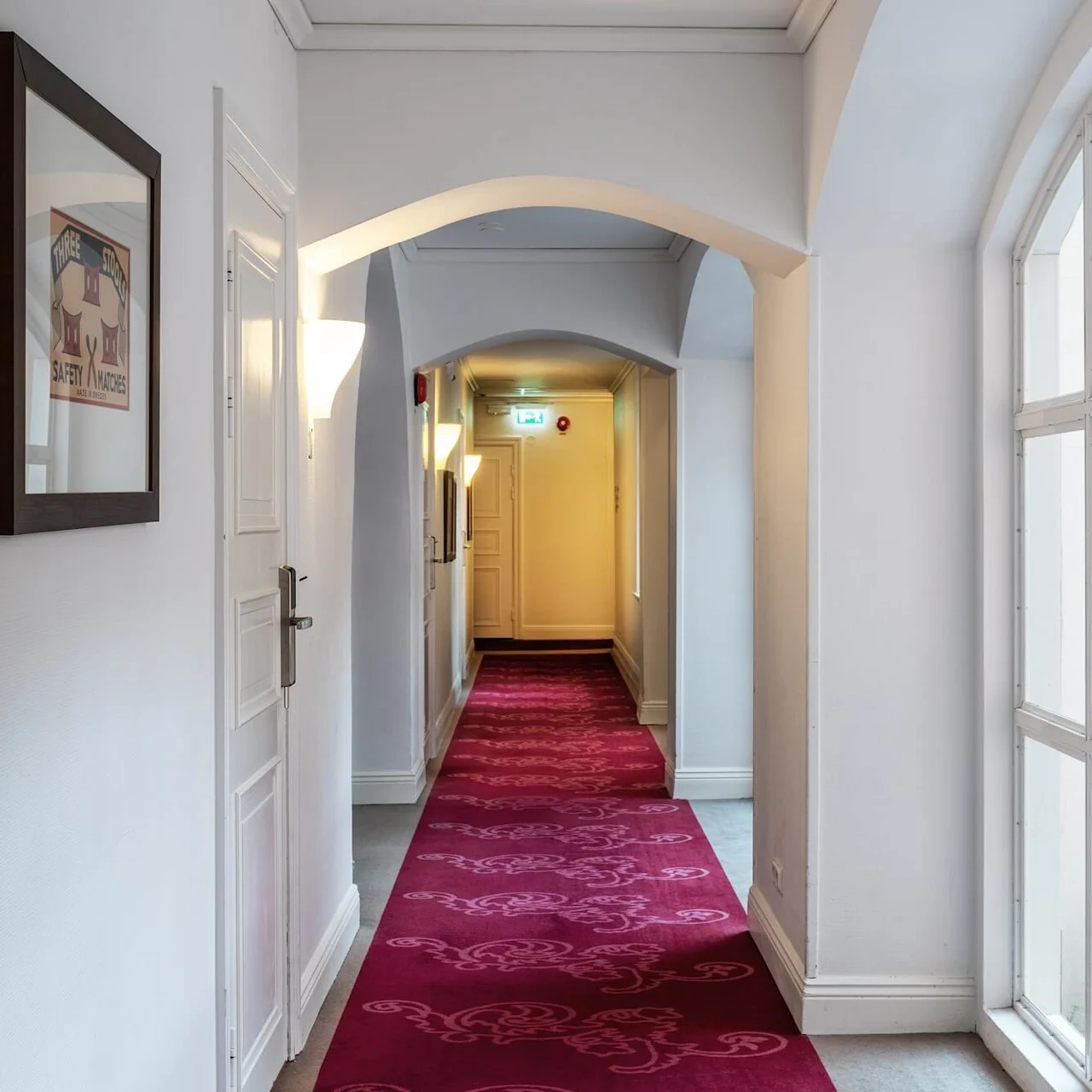 En tom hotelkorridor med röd matta, valvtak, inramat konstverk och livligt naturligt ljus genom stora fönster.