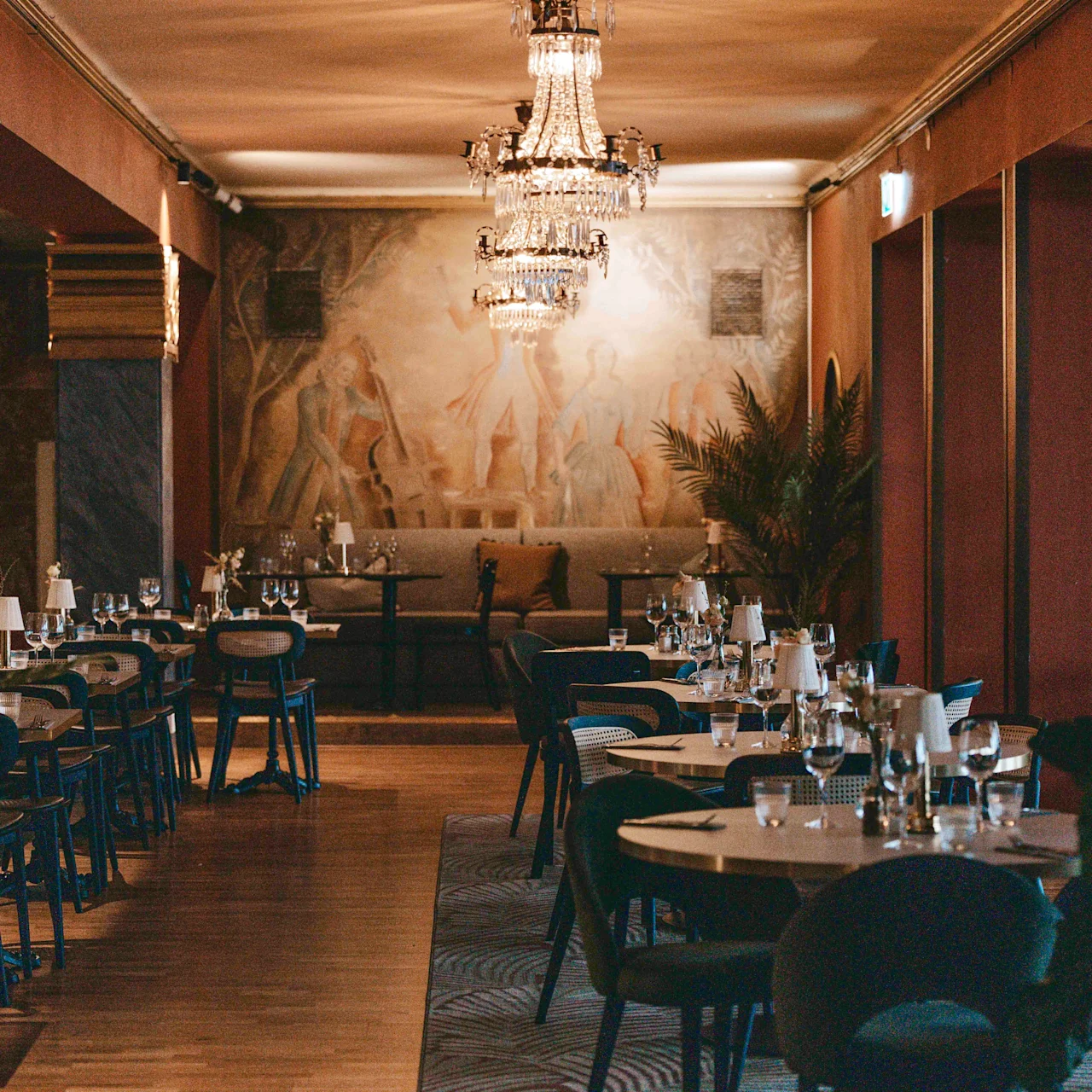 Tables set for dining; empty, adorned with glasses and cutlery. Action: Awaiting guests. Context: Elegant restaurant with chandeliers, wall art, and large plants, creating a sophisticated ambiance.