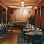 Tables set for dining; empty, adorned with glasses and cutlery. Action: Awaiting guests. Context: Elegant restaurant with chandeliers, wall art, and large plants, creating a sophisticated ambiance.