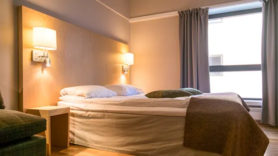 A neatly made bed with white linens and two pillows rests against a wooden headboard, flanked by wall-mounted lamps. A window with curtains provides natural light.