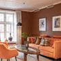 Oransje sofa og stol står i en koselig brunmalt stue, utsmykket med to innrammede bilder, en stålampe og et trebord, nær et vindu med lette gardiner.