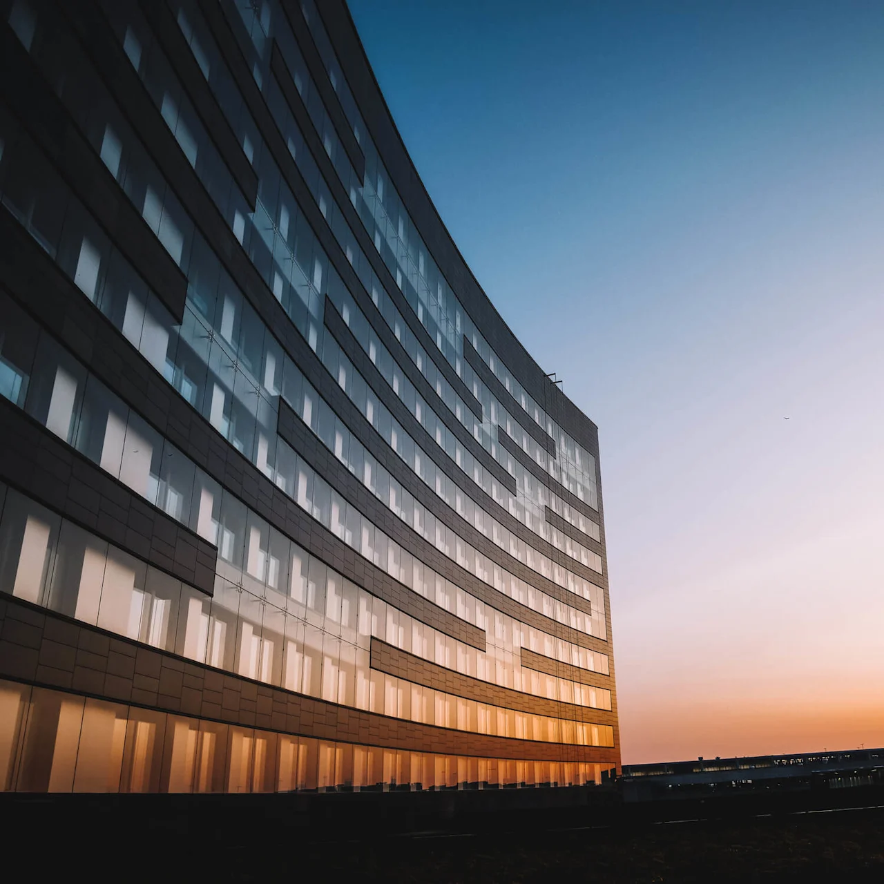 A curved glass building reflects a vibrant sunset, casting warm colors across its surface. The sky transitions from orange to blue, with city structures in the distant background.
