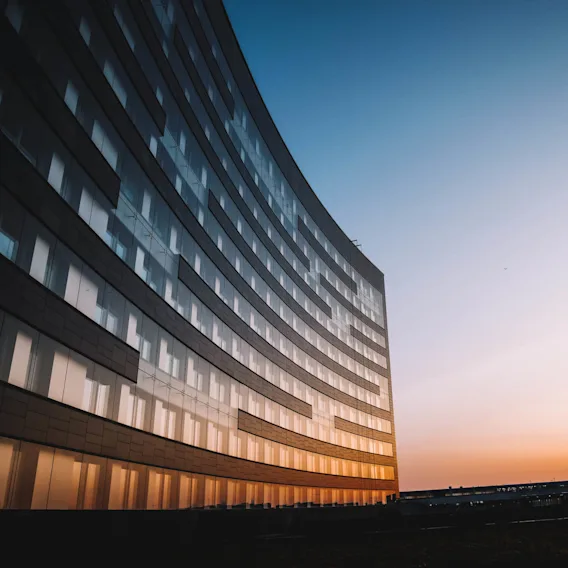 En böjd glasbyggnad reflekterar en livlig solnedgång, kastar varma färger över dess yta. Himlen övergår från orange till blå, med stadsstrukturer i bakgrunden.
